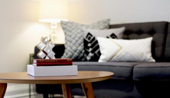 A living room with a wooden coffee table in the foreground with a stack of books on top. Sofa with throw pillows in the background with a side table and illuminated table lamp.